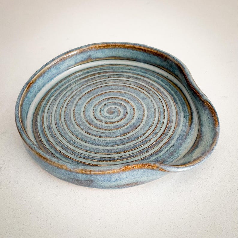 May include: A handmade ceramic spoon rest with a spiral design. The spoon rest is a light blue color with brown accents around the rim and spiral grooves. The rest has a curved edge for holding utensils.