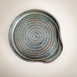 May include: A handmade ceramic spoon rest with a spiral design. The spoon rest is a muted blue with brown and gold accents. The rest has a curved indentation on one side.