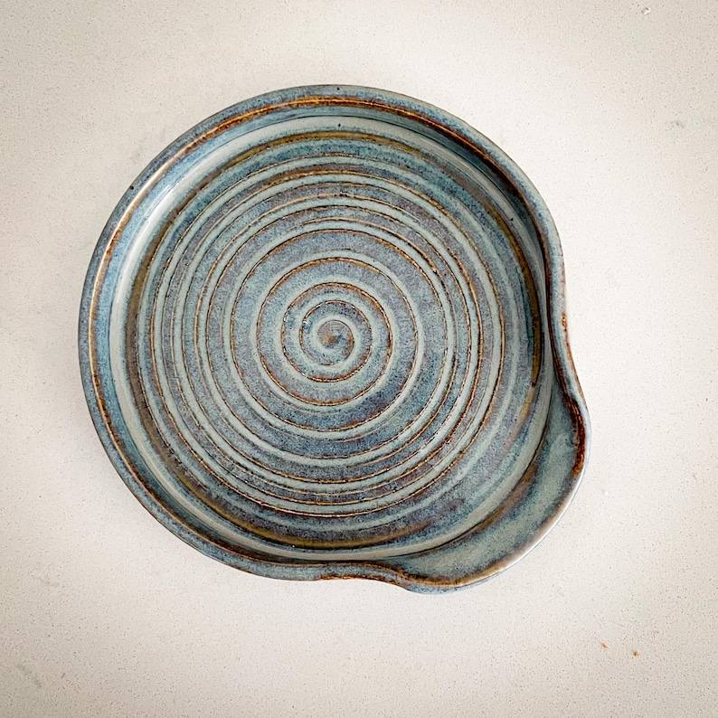 May include: A handmade ceramic spoon rest with a spiral design. The spoon rest is a muted blue with brown and gold accents. The rest has a curved indentation on one side.
