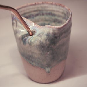May include: A handmade ceramic mug with a unique, organic shape. The mug features a two-tone glaze, with a light pink base and a flowing, marbled blue and white top. A copper-colored straw is inserted into a hole near the rim.