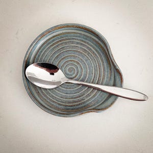 May include: A handmade ceramic spoon rest in a muted blue-green hue with a spiral design. A stainless steel spoon rests on the spoon rest. The spoon rest has a curved edge to hold the spoon. The spoon rest is a kitchen accessory.