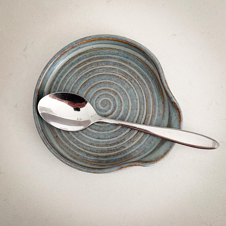May include: A handmade ceramic spoon rest in a muted blue-green hue with a spiral design. A stainless steel spoon rests on the spoon rest. The spoon rest has a curved edge to hold the spoon. The spoon rest is a kitchen accessory.