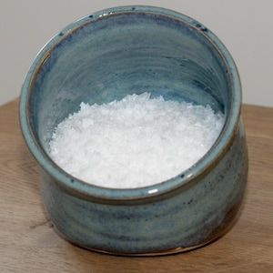 Puede incluir: Un salero de cerámica azul lleno de sal gruesa blanca. El salero tiene una forma redondeada con una abertura ligeramente inclinada. El exterior tiene un esmalte azul moteado con detalles marrones, sobre una superficie de madera.
