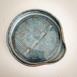 May include: A handmade, blue ceramic spoon rest with a wavy pattern in the center. The spoon rest has a rounded shape with a small lip on one side. The rim and the wavy pattern have a brown glaze.