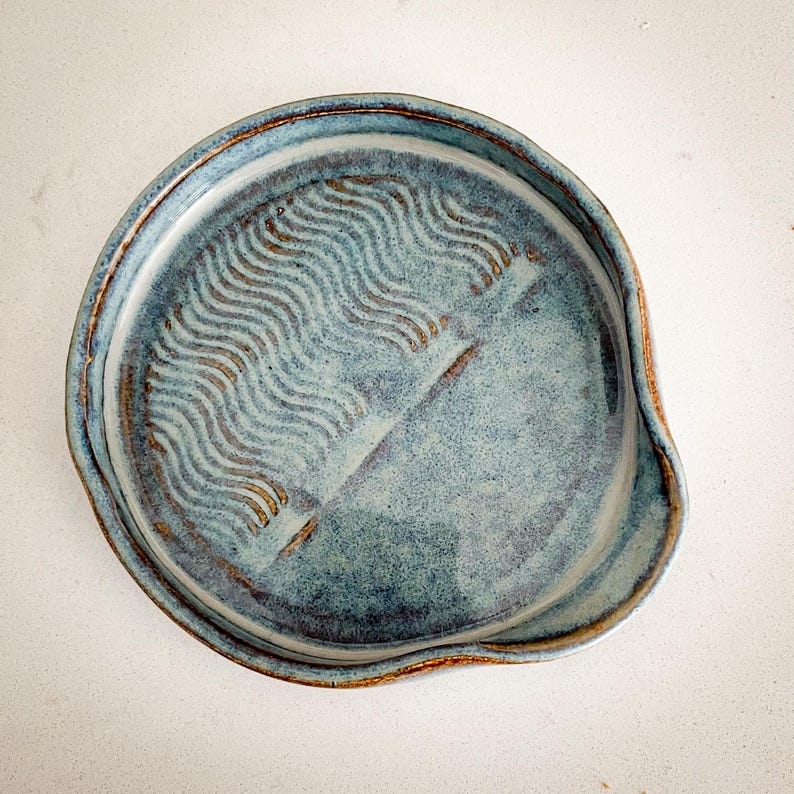 May include: A handmade, blue ceramic spoon rest with a wavy pattern in the center. The spoon rest has a rounded shape with a small lip on one side. The rim and the wavy pattern have a brown glaze.