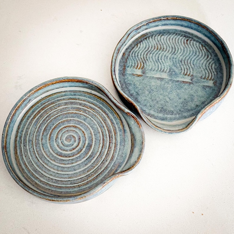 May include: Two handmade ceramic spoon rests in shades of blue and brown. One features a spiral design, the other has a wavy pattern. Both have a curved edge and a small spout for resting utensils.