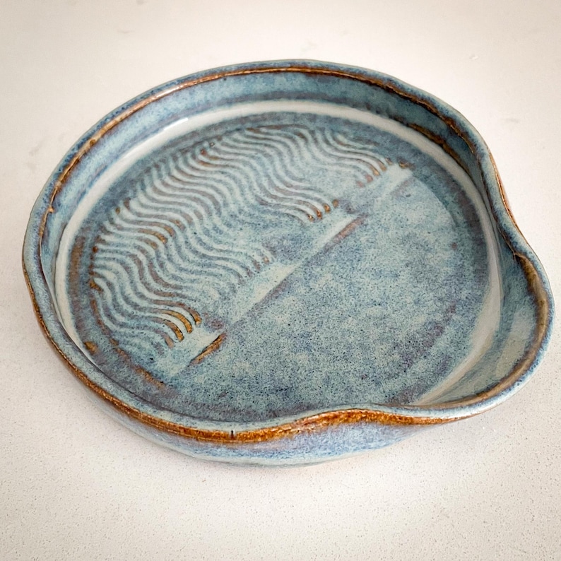 May include: A handmade ceramic spoon rest in a light blue glaze with a textured wave pattern. The rim and wave pattern have a brown accent. The spoon rest has a curved shape with a small lip to hold utensils.