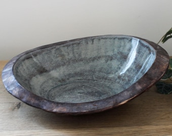 May include: A rustic, oval-shaped wooden bowl with a dark brown exterior and a muted teal interior. The bowl has a smooth, polished finish and sits on a wooden surface. The design is simple and natural, suitable for home decor.
