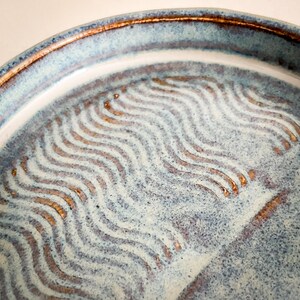 May include: Close-up of a handmade ceramic plate with a blue and white glaze. The plate features a wavy, textured pattern in a contrasting brown color. The rim has a darker brown border, adding a rustic touch to the design.