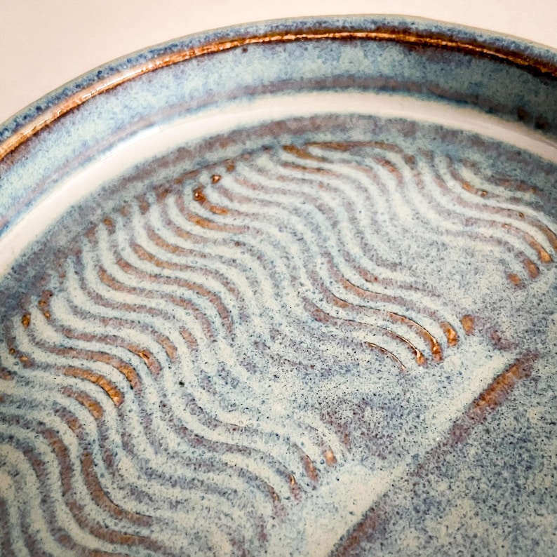 May include: Close-up of a handmade ceramic plate with a blue and white glaze. The plate features a wavy, textured pattern in a contrasting brown color. The rim has a darker brown border, adding a rustic touch to the design.