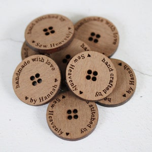 May include: A collection of round, wooden buttons with four holes each. The buttons are engraved with text that reads "handmade with love", "hand crafted by Hannah", and "Sew Heavenly". The buttons are a light brown color.