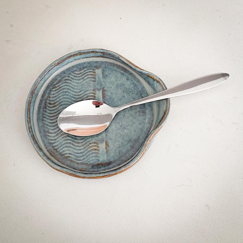 May include: A blue ceramic spoon rest with a wavy pattern and a stainless steel spoon. The spoon rest has a rounded shape with a lip to catch drips. The spoon is resting on the spoon rest.