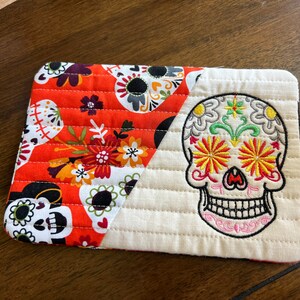 May include: A quilted orange and white fabric coaster with a colorful embroidered sugar skull design. The coaster features a variety of floral and skull patterns.