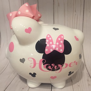 May include: White ceramic piggy bank with a pink bow, black Minnie Mouse ears with pink polka dots, and the name "Harper" written in pink. The piggy bank is decorated with pink and gray hearts.
