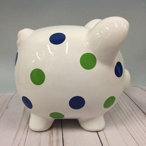May include: A white ceramic piggy bank decorated with blue and green polka dots. The piggy bank has four short legs, a small tail, and two ears. The coin slot is on top. The background is a light blue wall and a white wooden surface.