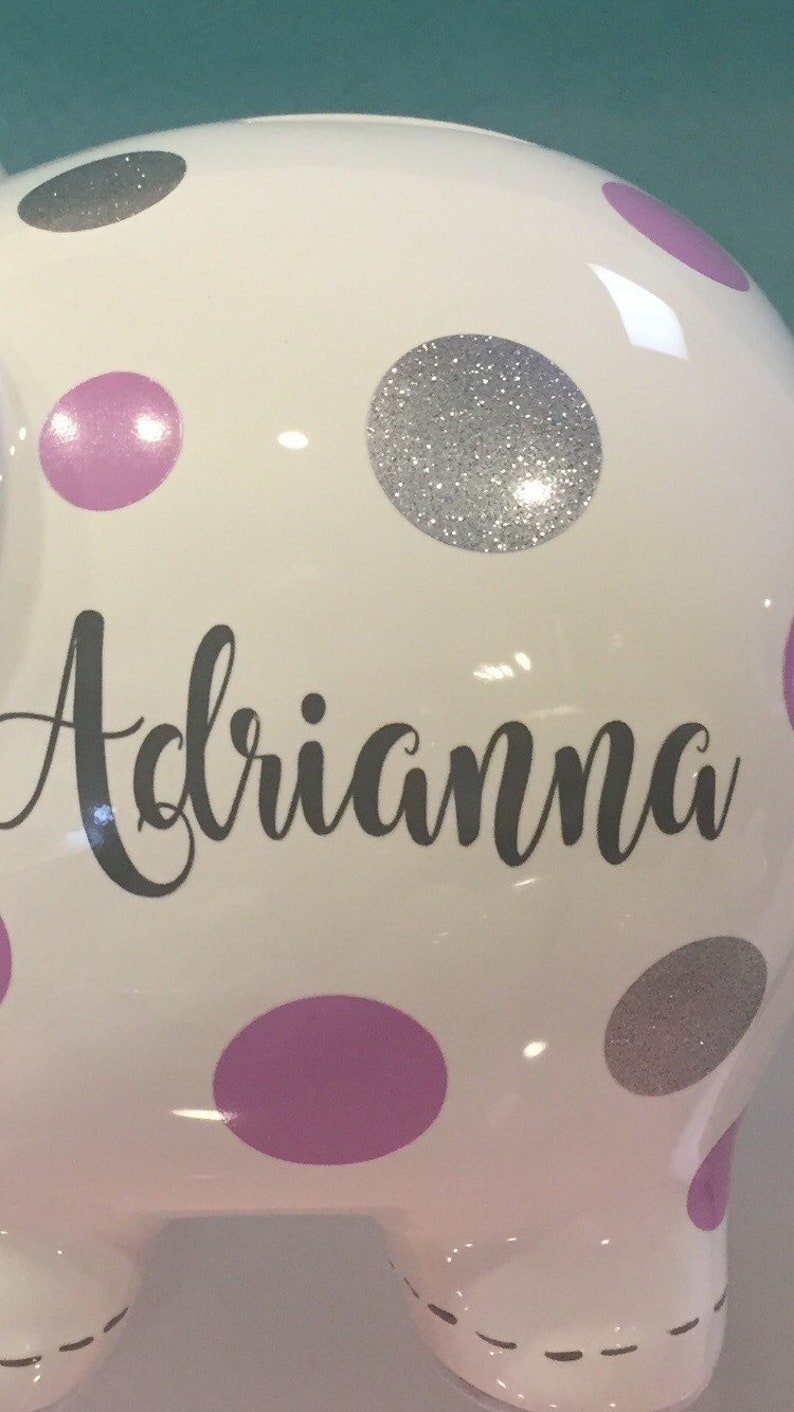 May include: White ceramic piggy bank with pink and silver polka dots. The name "Adriannna" is written in black cursive on the front.