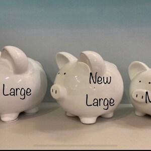 May include: Three white ceramic piggy banks of different sizes. The largest is labelled "Large", the middle one "New Large", and the smallest "Medium". The banks have small black eyes and nostrils.