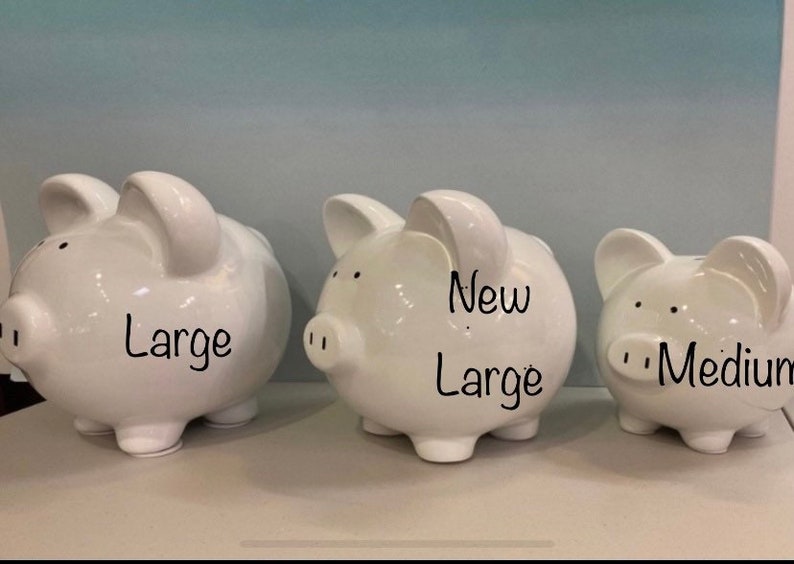 May include: Three white ceramic piggy banks of different sizes. The largest is labelled "Large", the middle one "New Large", and the smallest "Medium". The banks have small black eyes and nostrils.