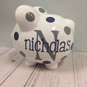 May include: White ceramic piggy bank with blue and grey polka dots. The piggy bank has the name "nicholas" and the letter "N" in blue and grey. The piggy bank is on a light wood surface.