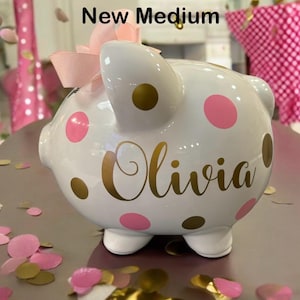 May include: White ceramic piggy bank with pink and gold polka dots. The piggy bank has the name "Olivia" written in gold lettering.