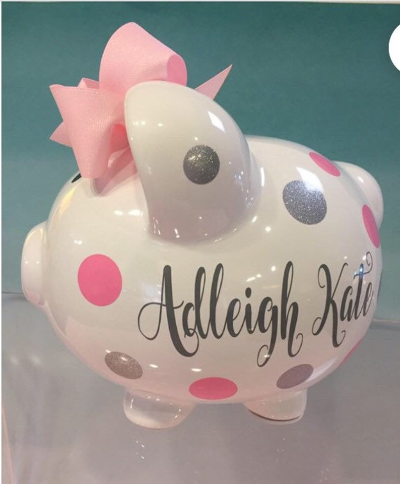 May include: White ceramic piggy bank with pink and silver glitter polka dots. The piggy bank has a pink bow on top and the name "Adleigh Kate" written on the side.