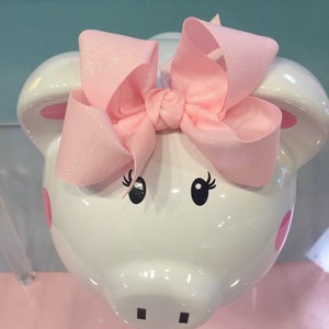 May include: White ceramic piggy bank with pink ears and a pink glitter bow on top. The piggy bank has black eyes with eyelashes and a pink nose.