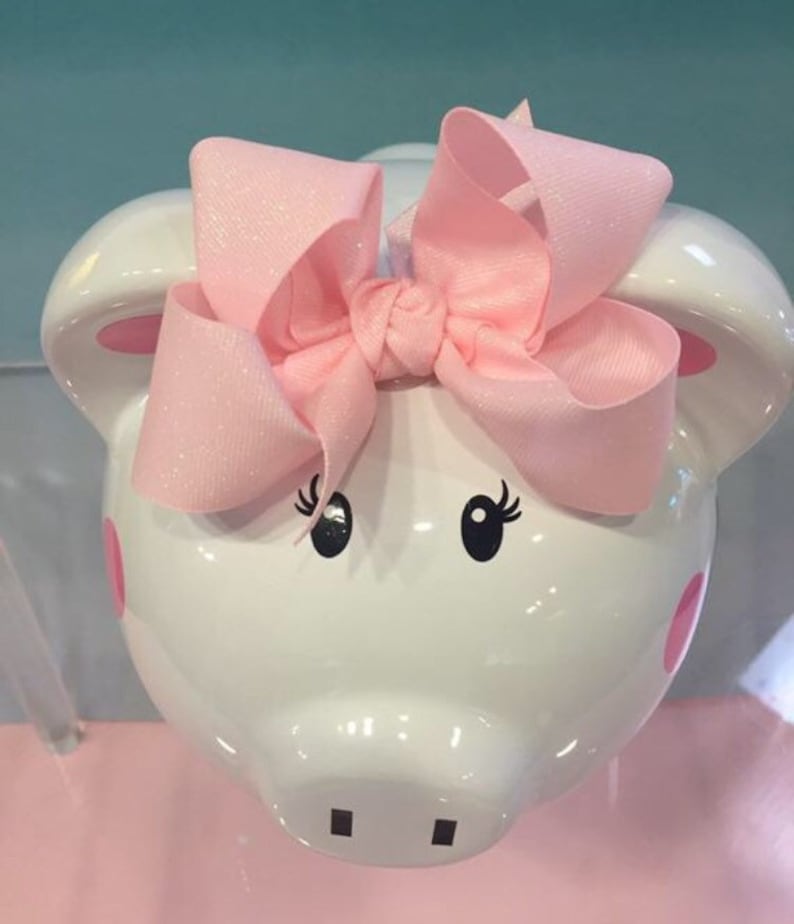 May include: White ceramic piggy bank with pink ears and a pink glitter bow on top. The piggy bank has black eyes with eyelashes and a pink nose.