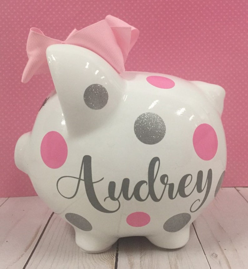 May include: White ceramic piggy bank with pink and silver glitter polka dots. The piggy bank has a pink bow on top and the name "Audrey" written in gray lettering.