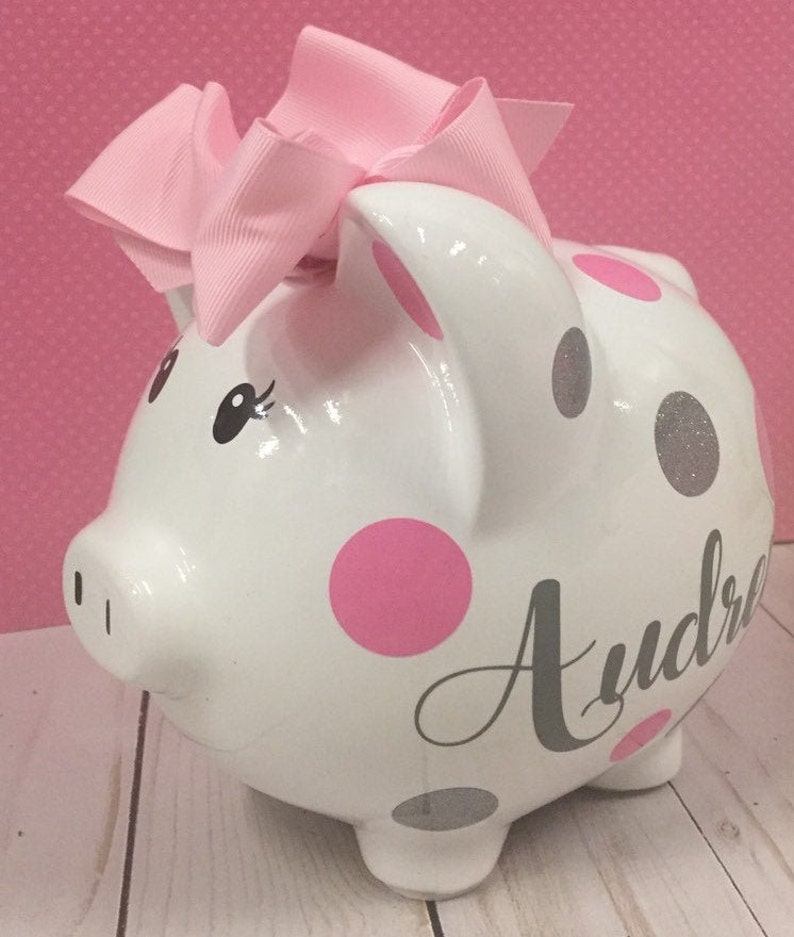 May include: White ceramic piggy bank with pink and silver polka dots. The piggy bank has a pink bow on top and the name "Audrey" written in gray cursive on the side.