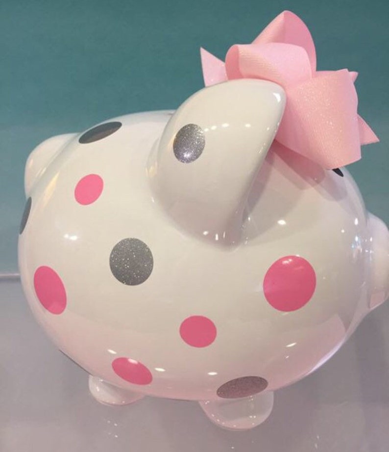 May include: White ceramic piggy bank with pink and silver glitter polka dots and a pink bow on top.