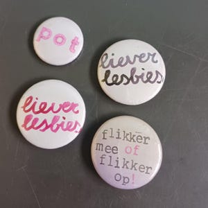 Op de afbeelding: Vier ronde witte knopen met verschillende berichten. De eerste knop zegt "pot" in roze letters. De tweede knop zegt "lieber lesbies" in zwarte letters. De derde knop zegt "lieber lesbies" in roze letters. De vierde knop zegt "flikker mee of flikker op!" in zwarte letters.