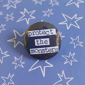 Könnte beinhalten: Ein runder Ansteckbutton mit dunklem Hintergrund und weißen rechteckigen Streifen, die den schwarzen Text "protect the monsters" in einer Schreibmaschinen-Schriftart zeigen. Das Design ist zentriert und klar und bietet eine kontrastreiche Optik.