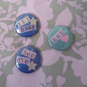 Könnte beinhalten: Drei runde, glitzernde Knöpfe mit Text und Stern- oder Herzakzenten. Zwei Knöpfe sind blau mit dem Text "zij haar" und "hen die" und einem Stern. Der dritte Knopf ist türkis mit einem rosa Herz und dem Text "het".
