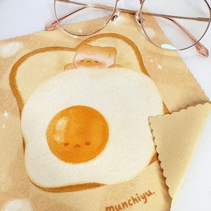Peut inclure: Un chiffon de nettoyage en microfibre jaune avec une illustration de dessin animé d'un œuf au plat sur du pain grillé. L'œuf a un visage mignon et le pain grillé a un petit chat endormi sur le dessus. Le texte "munchiyu." est imprimé dans le coin inférieur droit.