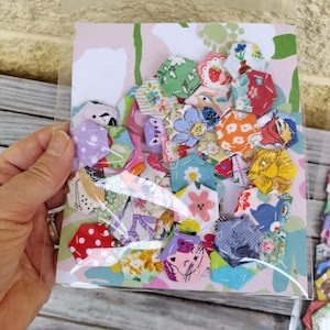 May include: A collection of small, fabric hexagon shapes featuring various colourful patterns, including floral, geometric, and animal prints, packaged in a clear plastic sleeve with a decorative backing card featuring a light pink and green floral design.
