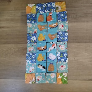 May include: A rectangular patchwork quilt composed of numerous square fabric patches. The design features a repeating pattern of various cat illustrations in orange, white, and black, interspersed with floral and botanical prints in blue, green, and yellow.