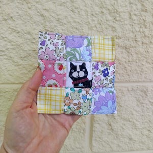 May include: A handmade patchwork coaster with a cat design. The coaster is made from fabric squares in floral and plaid patterns, including pink, yellow, and purple. The coaster is held in a hand. The coaster is approximately 9 cm x 9 cm.