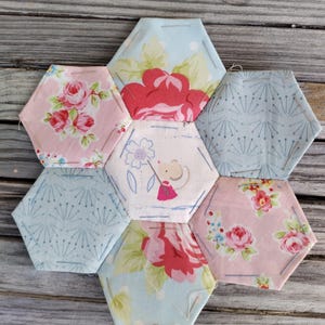 May include: A handmade patchwork coaster set featuring seven hexagonal pieces. The fabric pieces display floral patterns in pink and red, along with light blue and white designs. The coaster set is arranged on a wooden surface.