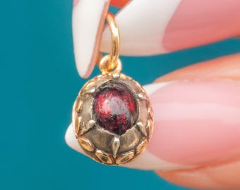 Puede incluir: Un colgante de oro con una piedra preciosa de color rojo oscuro en el centro. El colgante tiene forma ovalada con un borde dorado decorativo. La piedra preciosa es de color rojo oscuro y rico. El colgante está suspendido de un lazo dorado.