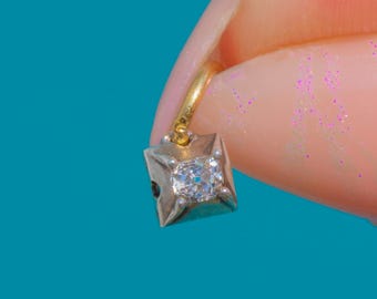 Puede incluir: Un pequeño colgante de forma cuadrada en tono plateado con una piedra transparente facetada en el centro, engastada en una montura de cuatro garras. El colgante está unido a una fina anilla circular dorada, creando una pieza compacta.