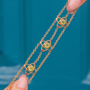 May include: A gold chain bracelet with three atom-shaped charms, each with a green gemstone in the center and small pink gemstones around the edges.