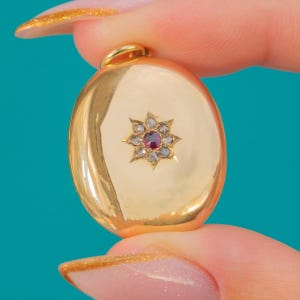 May include: A gold oval pendant with a starburst design. The starburst has a central red gemstone surrounded by smaller, clear stones. The pendant has a loop at the top for a chain.