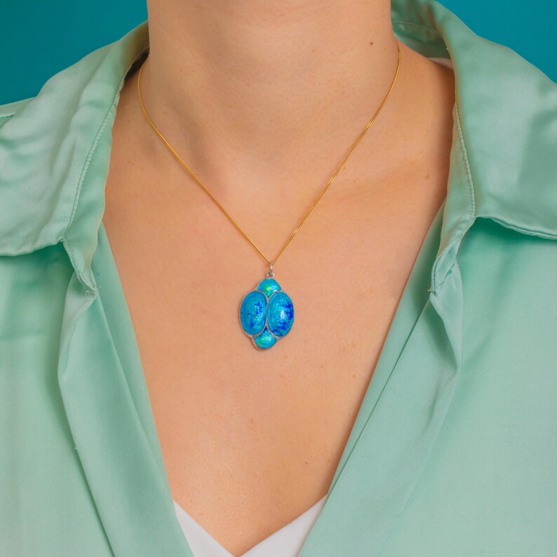 Puede incluir: Un collar dorado con un colgante que presenta un grupo de piedras azules similares al &oacute;palo. El colgante tiene un borde plateado. El collar se muestra sobre una camisa verde claro y un fondo turquesa.