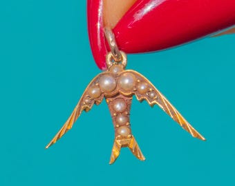 Puede incluir: Un colgante con forma de golondrina dorada que presenta una serie de pequeñas perlas redondas de color crema incrustadas a lo largo de las alas y el cuerpo central, con una pequeña anilla metálica en la parte superior.