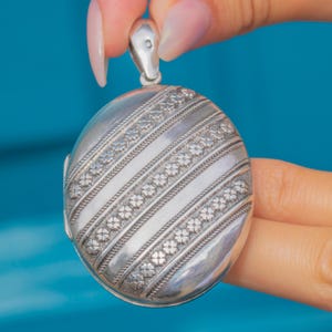 May include: Oval silver locket with diagonal striped design. The locket features floral and rope-like detailing. The top has a small loop for a chain. The background is a solid blue.