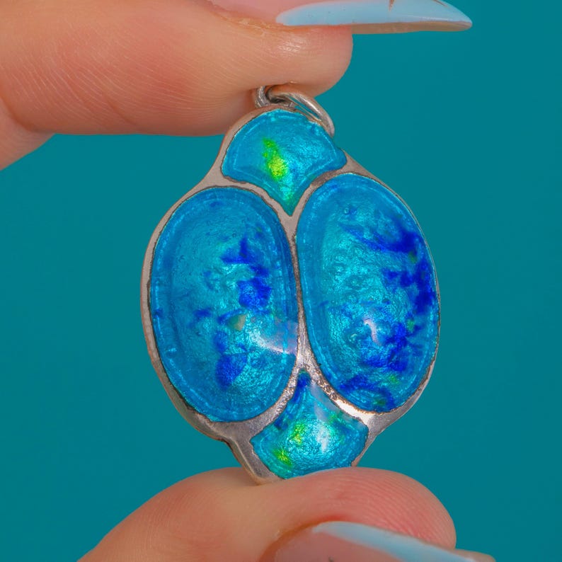 Puede incluir: Un colgante de plata con un dise&ntilde;o &uacute;nico. El colgante presenta una forma ovalada central con dos secciones de esmalte azul conectadas, acentuadas con detalles en azul y verde m&aacute;s claros. El colgante se sostiene entre dos dedos.