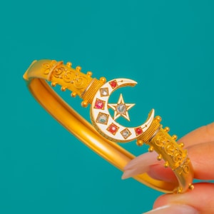 May include: A gold-toned hinged bangle bracelet featuring a central white crescent moon and star motif. The crescent is inlaid with alternating red and clear square-cut stones. The band has textured filigree detailing and small bead accents.