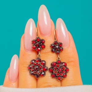 May include: A pair of drop earrings featuring two floral-shaped clusters of faceted deep red garnet stones set in gold-toned metal. Each earring consists of a smaller top flower and a larger bottom flower connected by a small metal link.