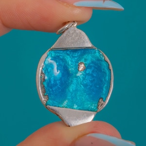 Puede incluir: Colgante de plata con centro de esmalte turquesa. El colgante tiene forma redondeada con una argolla de plata para una cadena. El esmalte tiene un aspecto texturizado con variaciones de color.