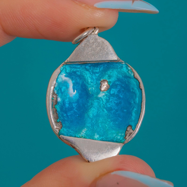 Puede incluir: Colgante de plata con centro de esmalte turquesa. El colgante tiene forma redondeada con una argolla de plata para una cadena. El esmalte tiene un aspecto texturizado con variaciones de color.